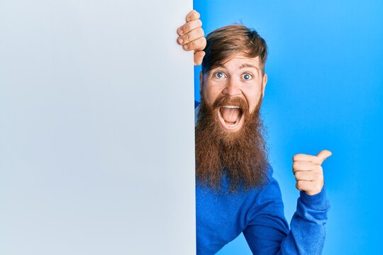 Young irish redhead man holding blank empty banner pointing thumb up to the side smiling happy with open mouth