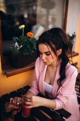 A brunette in pink clothes is sitting in a cafe with a glass of juice. Summer day.