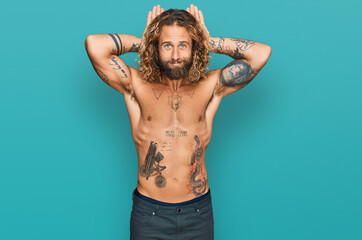 Handsome man with beard and long hair standing shirtless showing tattoos doing bunny ears gesture with hands palms looking cynical and skeptical. easter rabbit concept.
