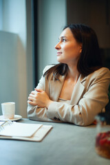 Young woman with long dark straight hair in in a beige nude business suit is talking on phone and writing notes