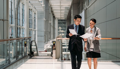 Fototapeta premium Two smiling colleagues walking and reading paper documents in office hall. Happy coworkers walking with papers and digital tablet in modern office building