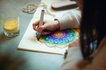 Young woman colouring mandala with markers and white rosary on table with cup of coffee at home