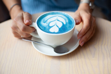 Unfocused woman hands holding cup of hot butterfly pea latte or blue spirulina latte on wooden table