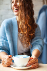 Unfocused happy woman with long wavy hair in warm sweater holding cup of hot blue latte