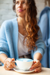 Unfocused happy woman with long wavy hair in warm sweater holding cup of hot blue latte