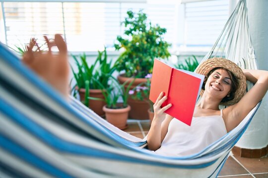Young beautiful caucasian woman smiling happy resting on a hammock reading a book at home