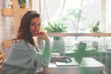 Young woman with long hair in blue knitted warm sweater sitting near the window and working on computer