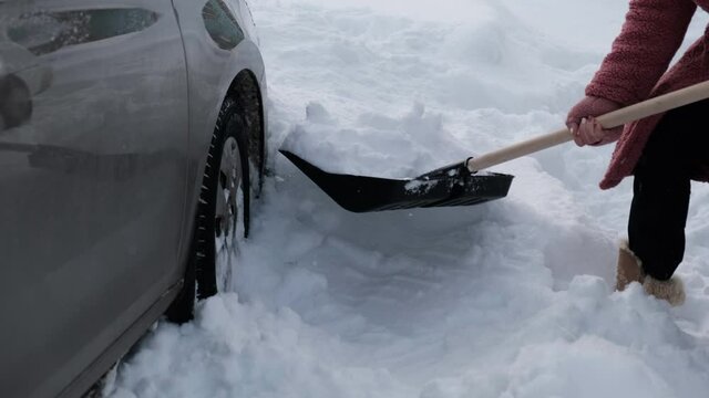 a young woman shovels snow away from her car after a heavy snowfall. the process of digging a car out of a snowdrift