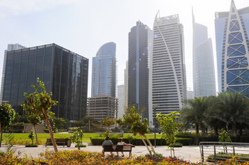 Vue sur un quartier résidentiel et un parc urbain au centre de Dubaï. Verdure, banc, personnes au premier plan et hauts bâtiments à l'architecture moderne en arrière-plan.