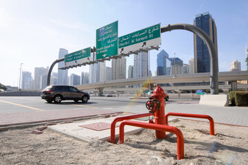 Vue urbaine sur une route à plusieurs voies, une arche de panneaux routiers et une voiture, avec une hydrante rouge au premier plan et les gratte-ciel de Dubaï en arrière plan.   