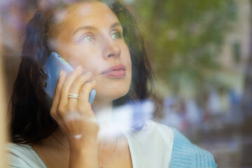 View through window of young woman talking on phone and working on computer with cup of hot coffee.