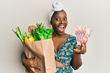 Young african woman holding groceries and 50 rand banknotes celebrating crazy and amazed for...
