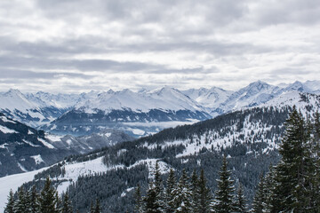 Fototapeta premium Behind the slope covered with coniferous forest, you can see the panorama of the Kitzbuhel Valley topped by a mountain range with sharp-pointed snow-capped peaks.