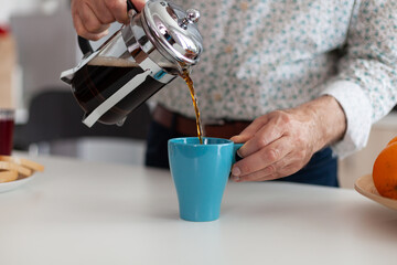 Fototapeta na wymiar Man's hand pouring aromatic coffee into a blue cup from french press on a wooden table during breakfast. Elderly person in the morning drinking fresh brown espresso from vintage mug, filter relax