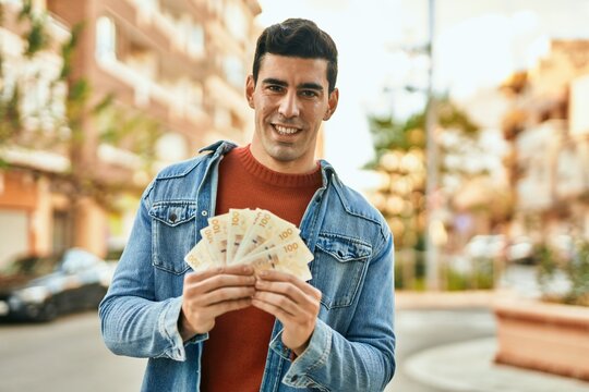 Young hispanic man smiling happy holding denmark krone banknotes at the city.