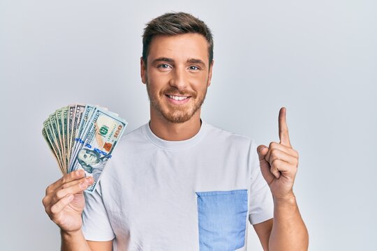 Handsome caucasian man holding dollars smiling with an idea or question pointing finger with happy face, number one