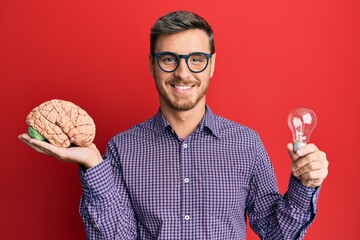 Handsome caucasian man holding brain and lightbulb for inspiration and idea smiling with a happy...