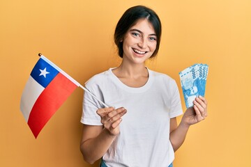 Young caucasian woman holding chile flag and chilean pesos banknotes smiling with a happy and cool smile on face. showing teeth.
