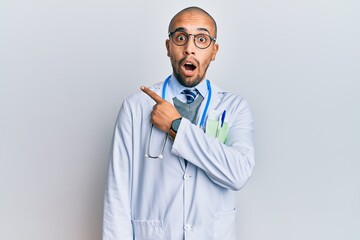 Hispanic adult man wearing doctor uniform and stethoscope surprised pointing with finger to the side, open mouth amazed expression.