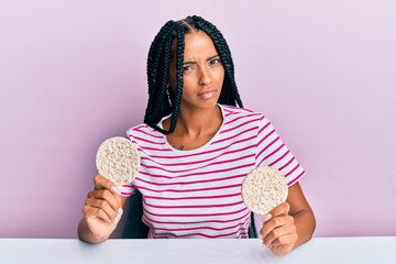 Beautiful hispanic woman eating healthy rice crackers in shock face, looking skeptical and sarcastic, surprised with open mouth