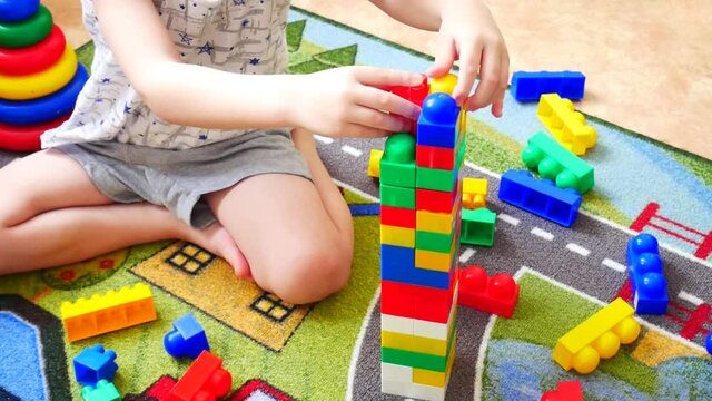 A little boy builds a tower using colorful constructor blocks sitting on the carpet close-up