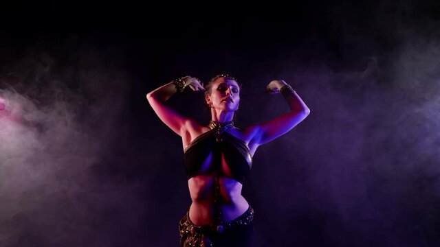 A woman dances an oriental dance on a dark smoky background in the studio, makes beautiful movements, holds a chain in her hands. She has an attractive body.
