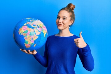 Young blonde woman holding world ball smiling happy and positive, thumb up doing excellent and approval sign