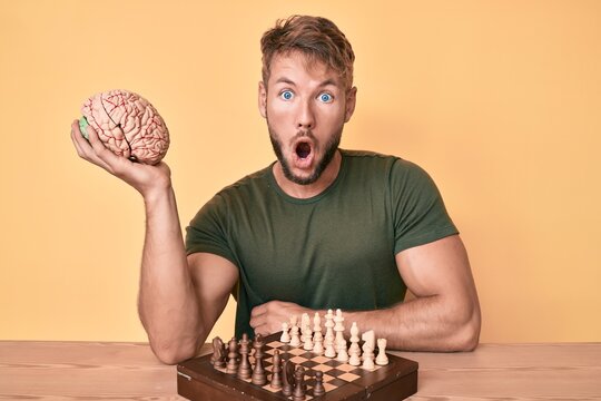 Young caucasian man holding brain while playing chess scared and amazed with open mouth for surprise, disbelief face
