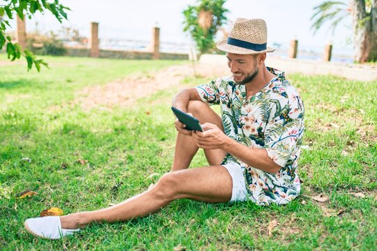 Caucasian handsome man smiling happy using touchpad device sitting on the grass at the park