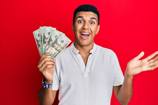 Young arab man holding dollars celebrating achievement with happy smile and winner expression with raised hand