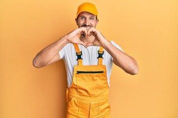 Young handsome man wearing handyman uniform over yellow background smiling in love doing heart symbol shape with hands. romantic concept.