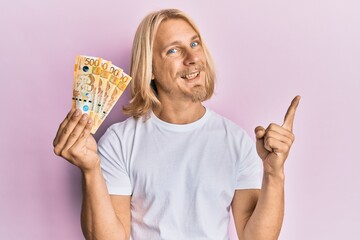 Caucasian young man with long hair holding 500 philippine peso banknotes smiling happy pointing with hand and finger to the side