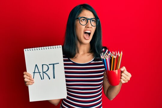 Beautiful asian young woman holding art notebook and colored pencils angry and mad screaming frustrated and furious, shouting with anger looking up.