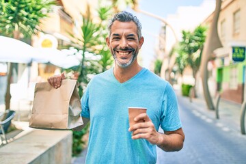 Middle age grey-haired man holding take away coffe and deliver paper bag with food at street of...