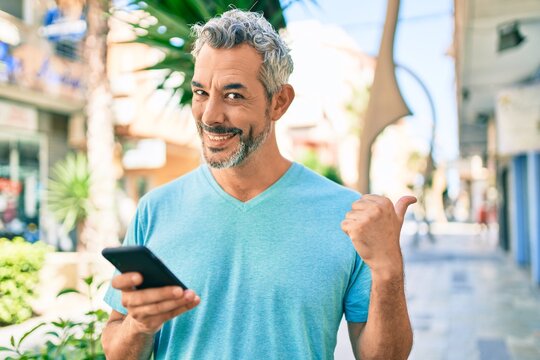 Middle age grey-haired man using smartphone at street of city pointing thumb up to the side smiling happy with open mouth