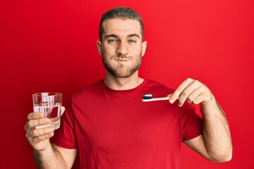 Young caucasian man holding toothbrush with toothpaste puffing cheeks with funny face. mouth inflated with air, catching air.