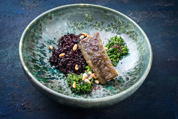 Modern style traditional fried skrei cod fish filet with baby broccoli, black rice and roasted pine nuts served as close-up on ceramic design plate with copy space