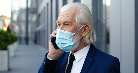 Close up of Caucasian old good-looking gray-haired man in medical mask talking on mobile phone outdoors. Retired senior rich businessman speaking on cellphone at city street. Conversation. Pandemic.