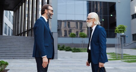 Caucasian senior businessmen in suits and ties meeting outdoor at business center, greeting with hands shaking and talking. Old rich men employees of office chatting. Business conversation in city.