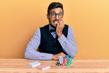 Handsome hispanic croupier man sitting on the table with poker chips and cards looking stressed and...