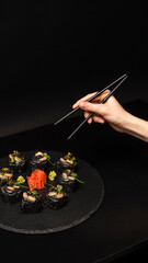 Hand with chopsticks wants to take custom sushi roll with black rice, crab meat, avocado, smoked salmon mousse, oar caviar, masago, shrimp cocktail, edible gold leaf, ginger, wasabi on black table.