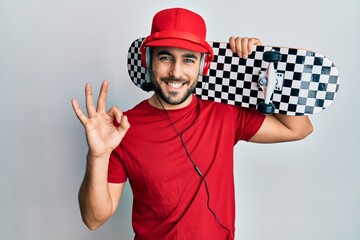 Young hispanic man using headphones holding skate doing ok sign with fingers, smiling friendly gesturing excellent symbol