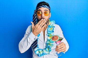 Young hispanic businessman wearing party funny style drinking cocktail covering mouth with hand,...