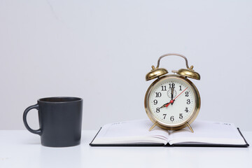 There is a vintage alarm clock on the open diary. There's a coffee cup next to it. The concept is...