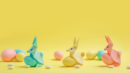 Paper Easter bunnies-origami made of colored paper and colored eggs on a delicate yellow background. The concept of the celebration of Easter, greeting card, crafts with your own hands.