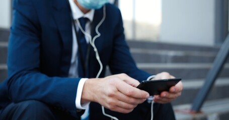 Caucasian middle-aged businessman in medical mask sitting on steps outdoor texting, tapping, playing or watching video on smartphone. Retired fired man in headphones listening to music on mobile phone