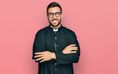 Young hispanic man wearing priest uniform happy face smiling with crossed arms looking at the...