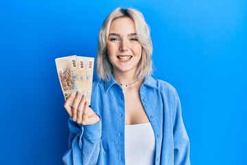 Young blonde girl holding new taiwan dollars banknotes looking positive and happy standing and smiling with a confident smile showing teeth