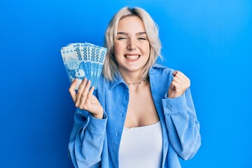 Young blonde girl holding 1000 chilean pesos screaming proud, celebrating victory and success very excited with raised arm