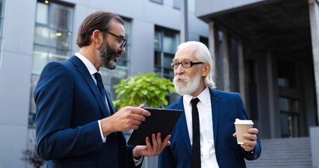 Caucasian senior businessmen standing at city street, drinking coffee and discussing information on tablet device. Serious men talking about work in morning, sipping drink. Business partners.